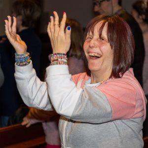 Include Choir Member smiling joyfully and holding hands up in the air Makaton signing