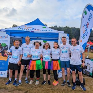 Photo of a team of 7 people standing side by side in a row at Run Reigate 2023 in Include.org tshirts standing in front of an Include.org stand or gazebo 