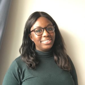 Image description: Folusho smiles at the camera. She has brown skin, shoulder length brown hair and glasses. She’s wearing a dark green turtleneck. End of image description.