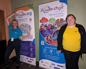 Charlotte an Include Choir Volunteer and Sarah and Include Choir Supported Volunteer stand at an event with some signs that say Include.org and Include Choir