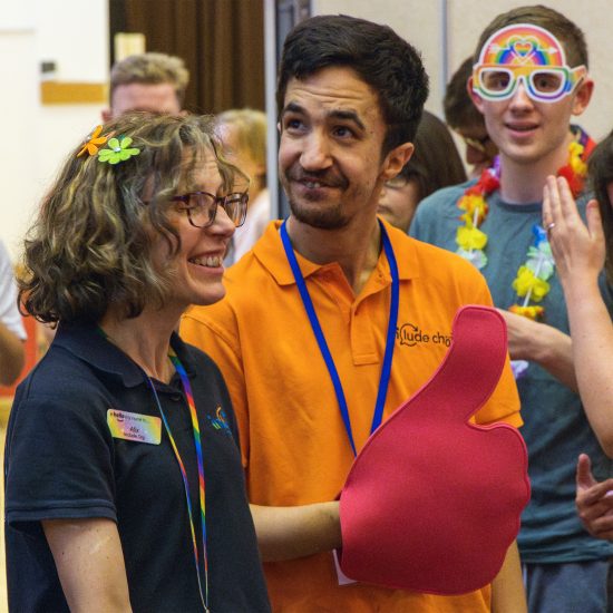 Alix Include's founder is pictured at and Include Choir rehearsal. A volunteer and a coir member are also in the photo. They are all looking in different directions and one person has a large glove on that looks like a thumbs up sign,