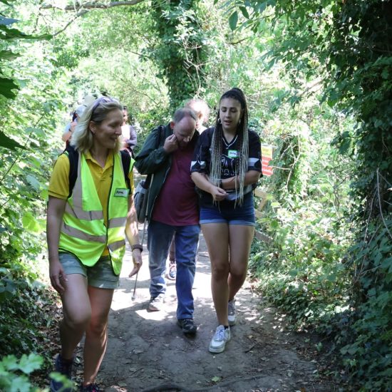 A photo of a group of people on a Stroll and Sign walk with Include. The photo shows a group leader Anna then a volunteer and a member all walking on a natural path in woodlands