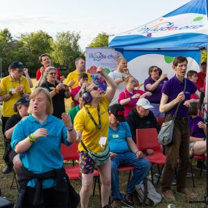 A small group of Include Choir members are standing near a tent in a park. They are singing and signing Makaton.