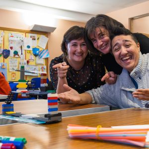 Three people from Include worked together as a team to build a bridge from lego. This is a photo of the three people proudly pointing at the Lego model bridge that they made without talking or writing words - as part of Include's bridge build challenge.