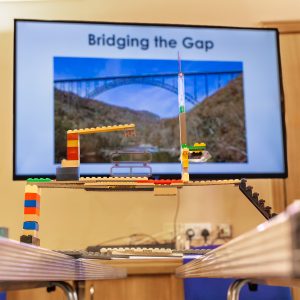 A photo of a bridge made from Lego that reaches over 30cm long across the gap between two tables. A screen in the background says 'Bridging the Gap' and shows a photo of a real bridge linking two land masses