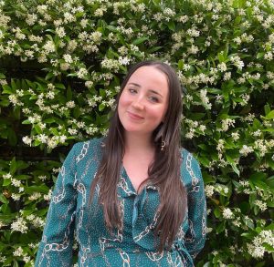 Eglantine was an intern at Include during the summer and this is a photo of her standing in front of a tall hedge and smiling.