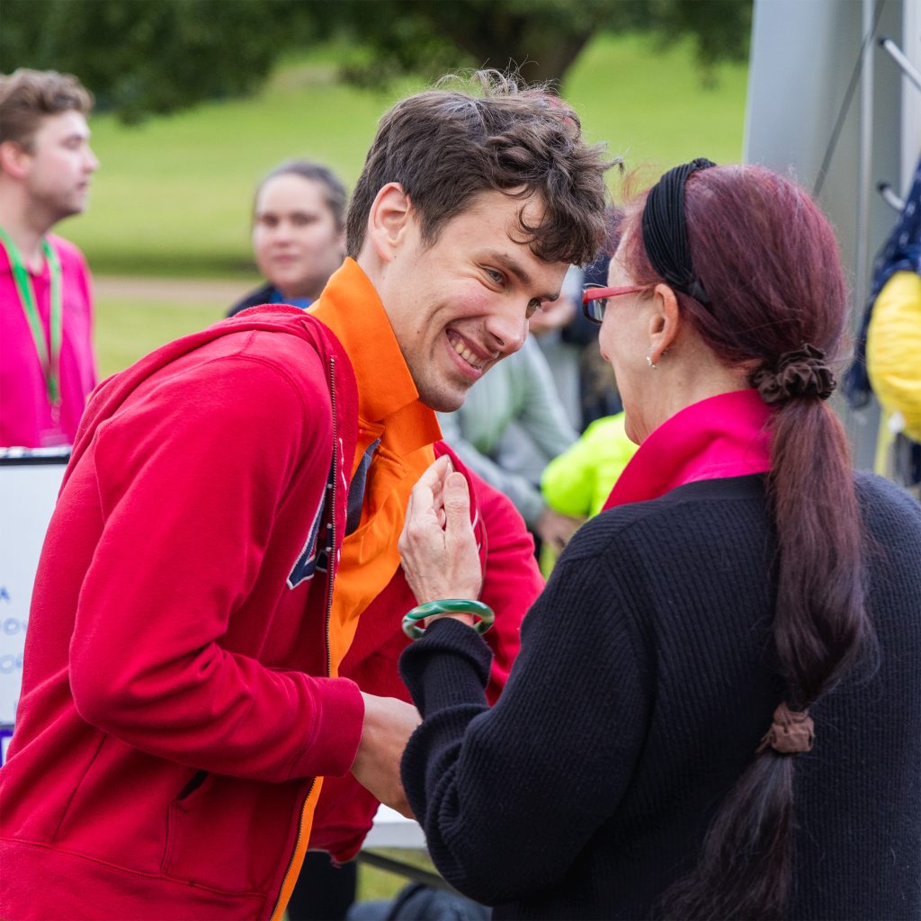 An outdoor shot of Include member Adam wearing a bright red zip-up hoodie over an orange shirt and is grinning broadly, at an Include Choir event.