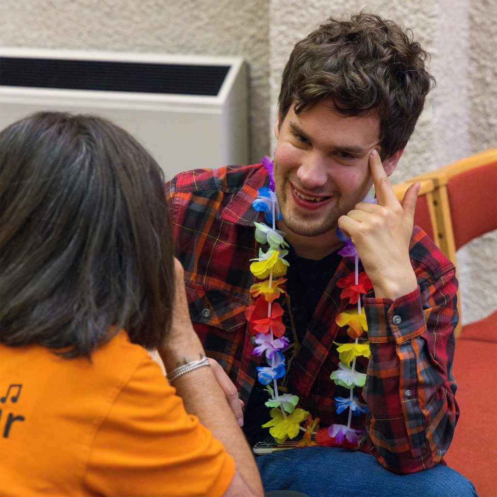 A close-up image of Include member Adam smiling and leaning forward while talking to Louise our volunteer coordinator. Adam is wearing a red and black plaid shirt and a bright rainbow coloured flower lei around his neck.
