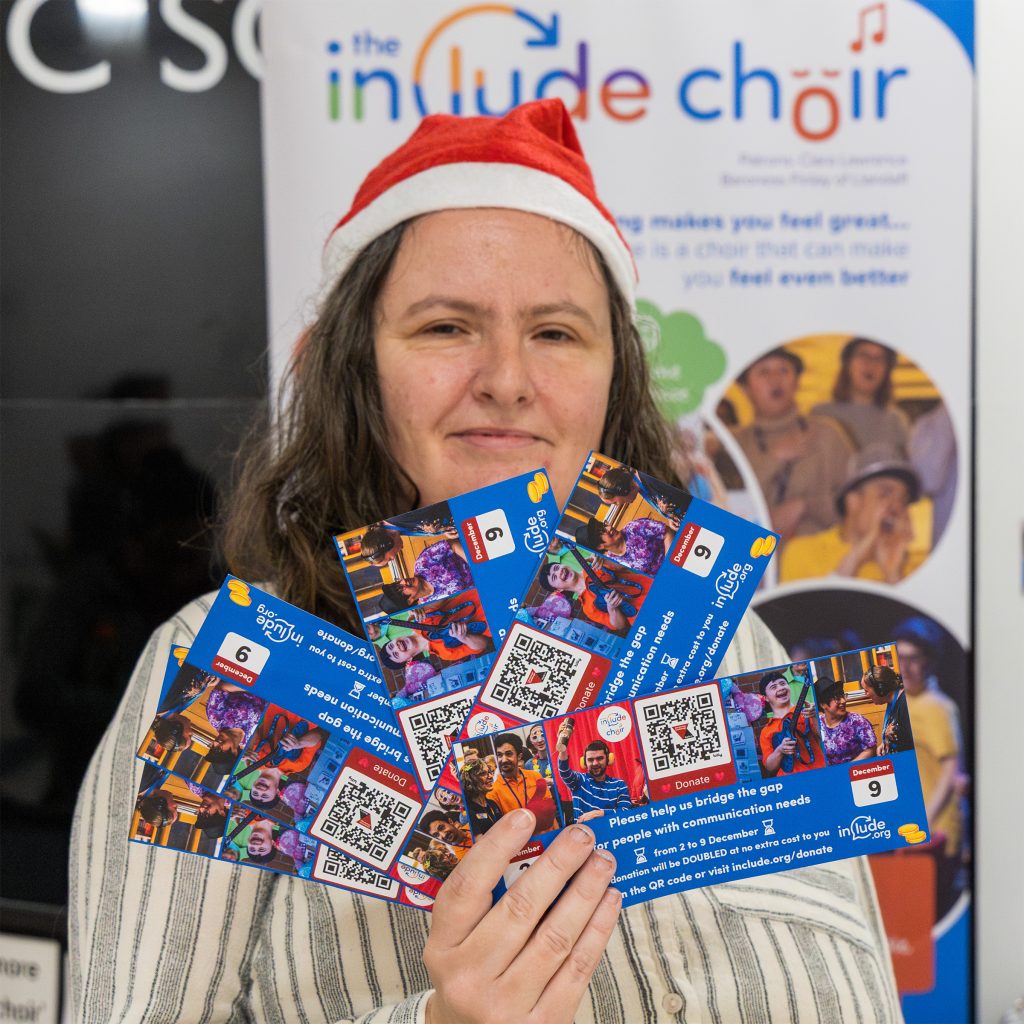 Include volunteer, Carol, wearing a Santa hat and holding a handful of Include Big Give fundraising leaflets to help raise money and share the Include.org message during a December community event in the Belfry shopping centre in Redhill.