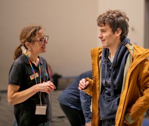 An image of Include member Adam, wearing a yellow hooded jacket over a navy hoodie. He is smiling and chatting with Alix, founder of Include.