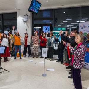 Include Choir members, staff and volunteers performing at a Christmas carol singing event at Waitrose in Banstead. The group is using Makaton signs while singing for the community.