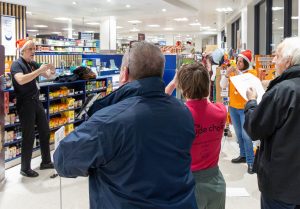 Include Choir members, staff and volunteers performing at a Christmas carol singing event at Waitrose in Banstead. The group is using Makaton signs while singing for the community.