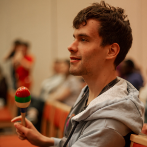 A side profile of Include member Adam sitting in a chair at a Redhill Choir session, wearing a light blue zip-up hoodie. He holds a colourful maraca with red, green, and white stripes.